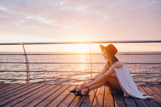 Femme En Robe Blanche Assise Au Bord De La Mer Au Lever Du Soleil Dans Une Ambiance Romantique Portant Un Chapeau Rouge