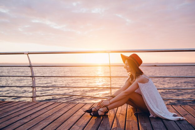 Femme en robe blanche assise au bord de la mer au lever du soleil dans une ambiance romantique portant un chapeau rouge
