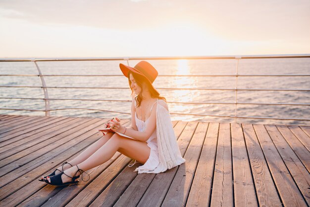 Femme en robe blanche assise au bord de la mer au lever du soleil en pensant et en prenant des notes dans le livre de journal