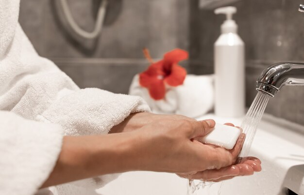 Une femme en robe de chambre se lave les mains avec du savon sous l'eau courante d'un robinet.