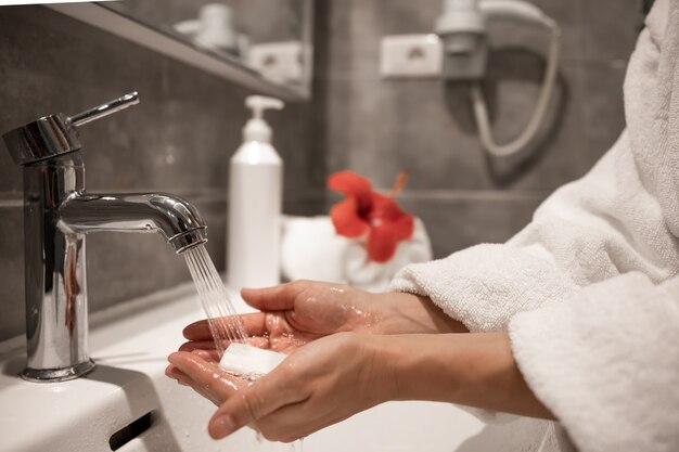 Une femme en robe de chambre se lave les mains avec du savon sous l'eau courante d'un robinet