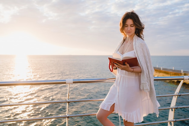 Photo gratuite femme en robe d'été blanche marchant au bord de la mer au lever du soleil avec livre de journal dans la pensée de l'humeur romantique et prendre des notes