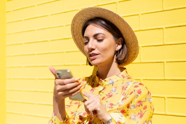 Femme en robe d'été jaune et chapeau sur mur de brique jaune calme et positif holding mobile phone