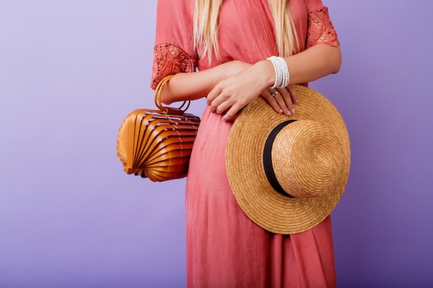 femme en robe rose tendance et chapeau de paille tenant un sac en bambou sur violet.