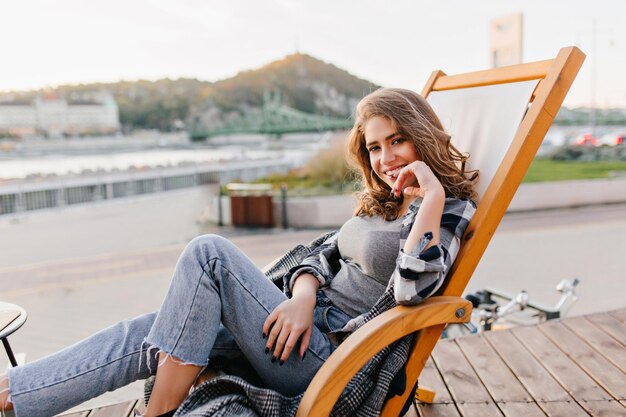 Femme romantique en jeans se reposant dans un fauteuil inclinable et touchant son visage de manière ludique. Portrait en plein air d'une fille bouclée en chaise longue posant sur fond de nature par temps froid.