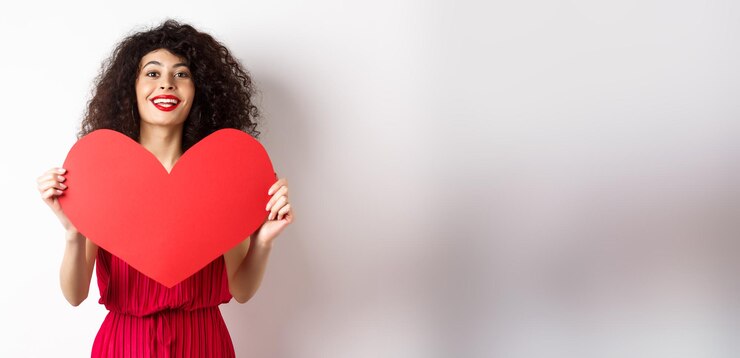 Photo gratuit femme romantique en robe montrant un grand coeur rouge tombant amoureux souriant heureux à l'arrière-plan blanc de l'appareil photo
