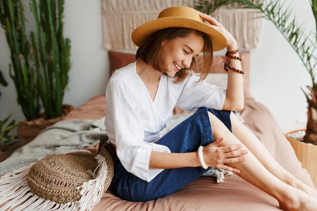 Femme romantique avec un sourire candide assis sur le lit, profitant d'une matinée ensoleillée dans son appartement élégant dans un style bohème