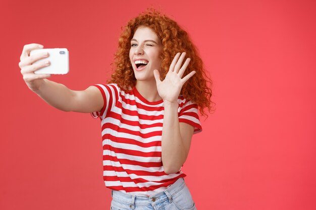 Une femme rousse aux cheveux bouclés, joyeuse et pleine d'énergie, envoie un bonjour en agitant la paume souriante ...