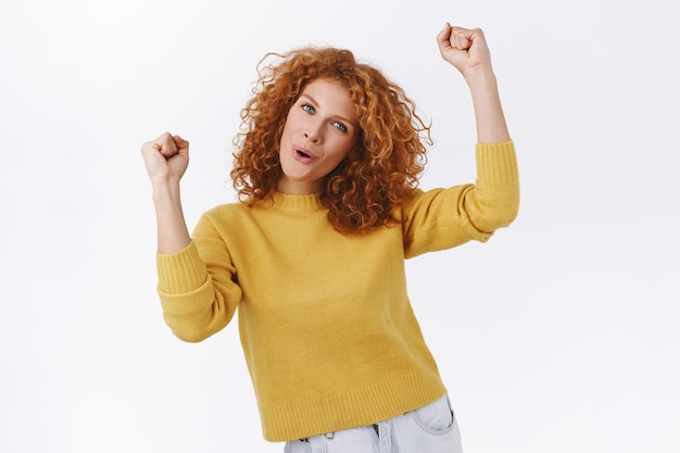 femme rousse aux cheveux roux bouclés, vêtue d'un pull jaune, levant les mains sur blanc