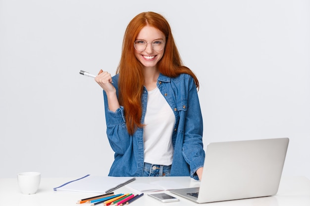 Une femme rousse charismatique séduisante et confiante près d'un ordinateur portable et d'une table avec des fournitures de dessin, une caméra souriante et mystique, un fond blanc debout a un excellent plan.