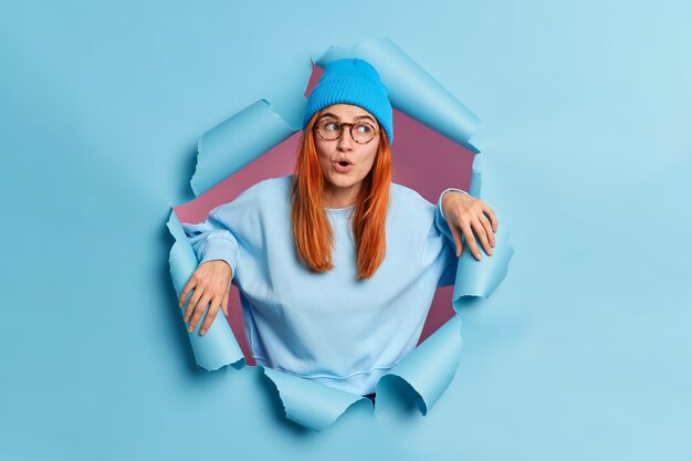 La femme rousse choquée garde la bouche ouverte de l'émerveillement regarde ailleurs avec étonnement porte un chapeau et un pull à manches longues.