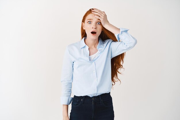 Une femme rousse choquée et inquiète haletant et regardant devant elle, effrayée, gifle le front comme oubliant quelque chose, debout sur un mur blanc alarmée