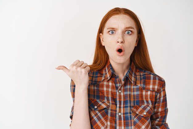 Femme rousse choquée à la peau pâle et sans maquillage pointant du doigt la bannière haletant étonnée devant la caméra debout sur fond blanc