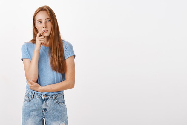 Femme rousse concentrée et curieuse réfléchie en t-shirt bleu et jeans mordant le doigt intrigué à droite tout en pensant avoir le désir ou l'intérêt pour quelque chose