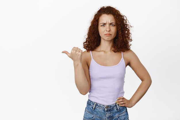 Femme rousse confuse et mécontente, fronçant les sourcils, pointant vers la gauche avec un visage en colère interrogé, n'aimant pas qch, debout sur fond blanc.