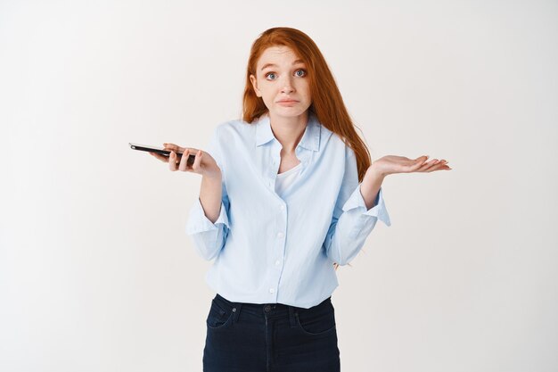 Femme rousse confuse tenant un téléphone et haussant les épaules, l'air désemparée à l'avant, mur blanc