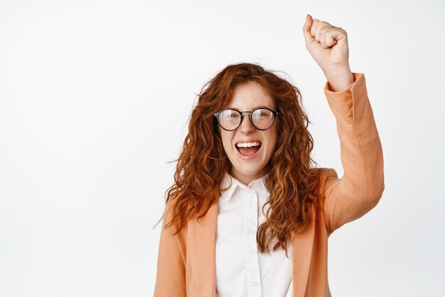 Femme rousse encouragée acclamant levant le poing et criant en chantant ou enracinant pour quelqu'un qui proteste debout heureux sur fond blanc