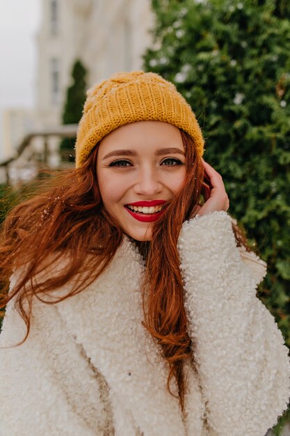 Femme rousse excitée en bonnet tricoté posant en hiver. Dame de gingembre romantique en manteau souriant en plein air.