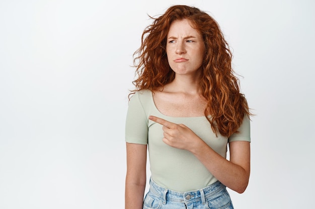 Photo gratuite femme rousse grincheuse pointant et regardant à gauche avec l'expression du visage moue portant un t-shirt et un jean fond blanc