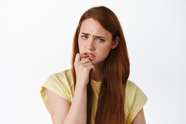 Une femme rousse hésitante a des doutes, touchant la lèvre et fronçant les sourcils, regardant la caméra et réfléchissant, pensant à quelque chose de sérieux, fond blanc