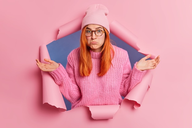 Une femme rousse inconsciente et perplexe étend les paumes avec une expression désemparée qui porte les lèvres et ne sait pas quoi faire porte un chapeau rose à la mode et un pull en tricot traverse le papier