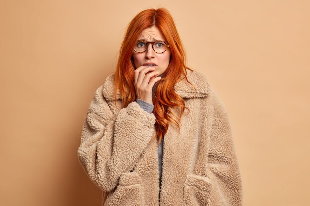 Une femme rousse inquiète mécontente mord les ongles des doigts semble anxieuse vêtue d'un manteau de fourrure chaud.