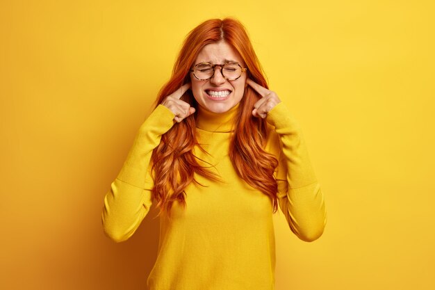 Une femme rousse insatisfaite serre les dents et se bouche les oreilles agacée par un son ou un bruit fort porte un pull décontracté.