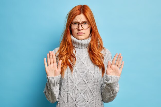 Une femme rousse mécontente grave montre un geste d'arrêt lève les paumes vers un pull gris en tricot.