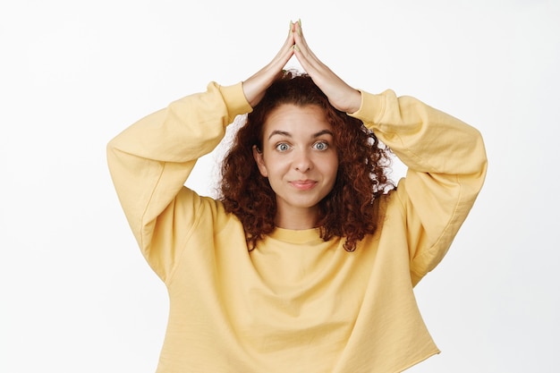 Femme rousse mignonne et idiote faisant un geste sur le toit d'une petite maison au-dessus de la tête, toit à la main sur le dessus et souriant, debout sur blanc.