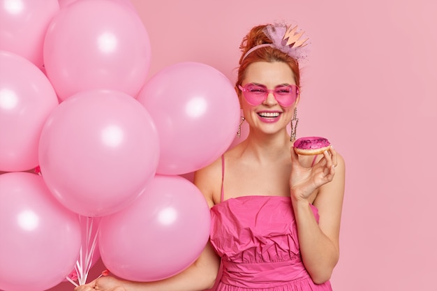 Une femme rousse à la mode positive sourit largement a une humeur festive tient de délicieux ballons gonflés de beignets porte des lunettes de soleil et une robe en forme de coeur