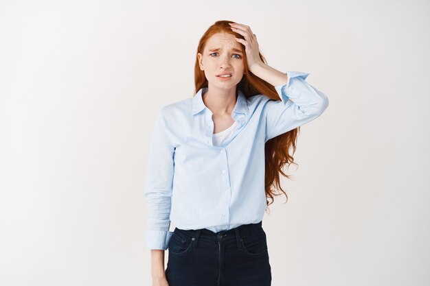 Femme rousse nerveuse à l'air inquiète et bouleversée, touchant la tête et regardant devant coupable, a oublié quelque chose, debout sur un mur blanc