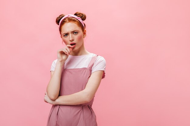 Femme rousse avec des petits pains regarde pensivement la caméra. Portrait de femme en combinaison rose et haut blanc.