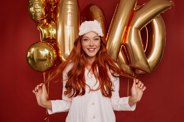 femme rousse en pijama et masque de sommeil posant excité avec des ballons dorés sur rouge