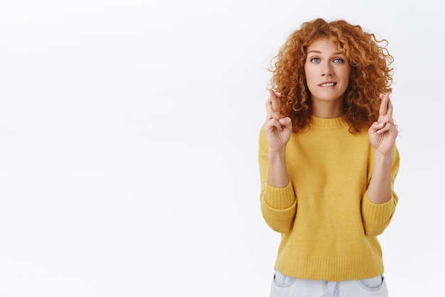 Une femme rousse pleine d'espoir et d'insécurité, inquiète, aux cheveux bouclés, porte un chandail jaune, croise les doigts, bonne chance, mord la lèvre nerveusement et regarde la caméra anxieuse en anticipant le rêve devenu réalité en attendant le résultat