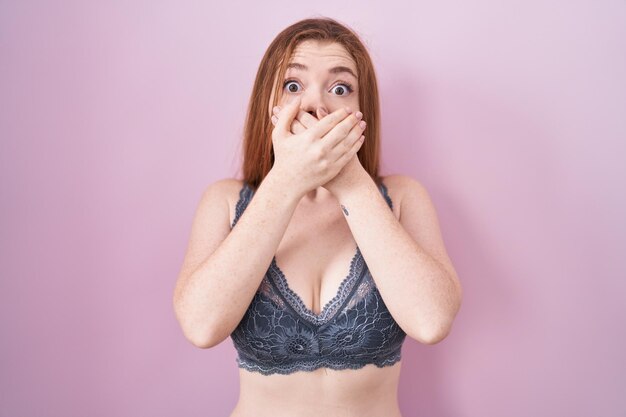 Femme rousse portant de la lingerie sur fond rose choquée couvrant la bouche avec les mains pour erreur. notion secrète.