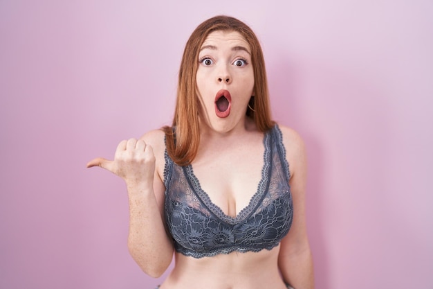 Femme rousse portant de la lingerie sur fond rose surprise pointant avec le doigt de la main sur le côté, bouche ouverte expression étonnée.