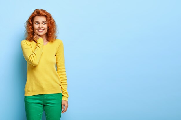 Une femme rousse positive a de la bonne humeur, garde la main sur le cou, a un regard joyeux de côté, porte une tenue jaune et verte