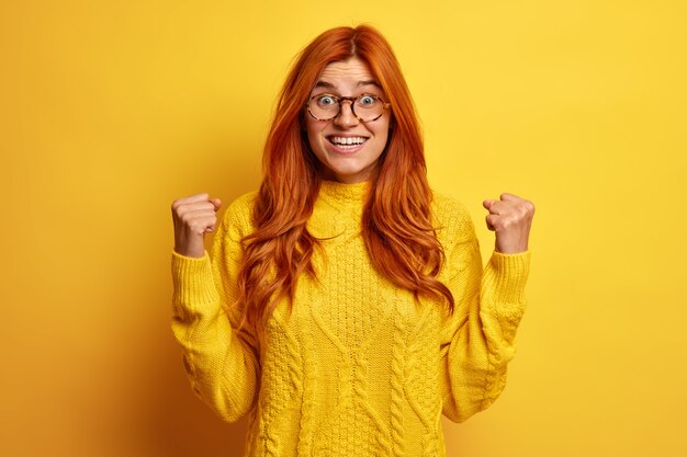 Une femme rousse ravie euphorique lève les poings fermés fait un geste oui excité par d'excellentes nouvelles porte des lunettes et un pull jaune célèbre l'obtention de prix à l'intérieur. Concept de victoire de succès