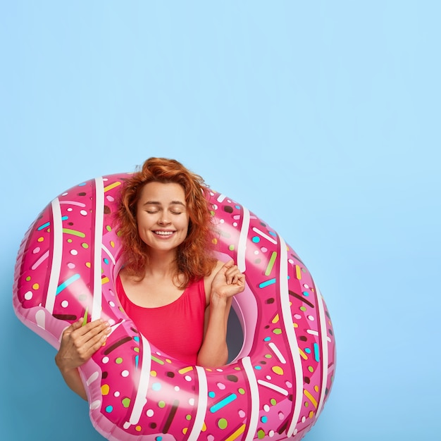 Une femme rousse ravie et ravie serre le poing de satisfaction, anticipe l'heure d'été, garde les yeux fermés, se tient avec une bague gonflée, se tient contre un mur bleu avec un espace de copie. Les gens et le repos