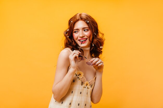 Femme rousse en robe à carreaux sourit et mord la manille de lunettes de soleil sur fond jaune.