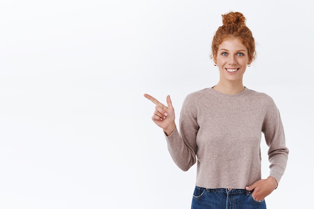 Femme rousse séduisante et charismatique aux cheveux bouclés peignés en chignon désordonné, tenir la main dans la poche, pointant vers la gauche, présenter le produit, souriant satisfait comme cadeau publicitaire, mur blanc debout