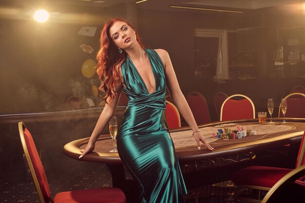 Une femme rousse sexy vêtue d'une longue robe en satin bleu pose assise sur une table de poker et regarde la caméra dans un casino de luxe. Passion, cartes, jetons, alcool, victoire, jeu - c'est comme divertissement féminin