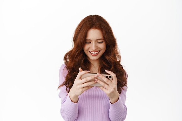 Femme rousse souriante regardant une vidéo sur l'écran d'un smartphone, regardant un téléphone portable heureux, jouant à un jeu vidéo, debout sur fond blanc