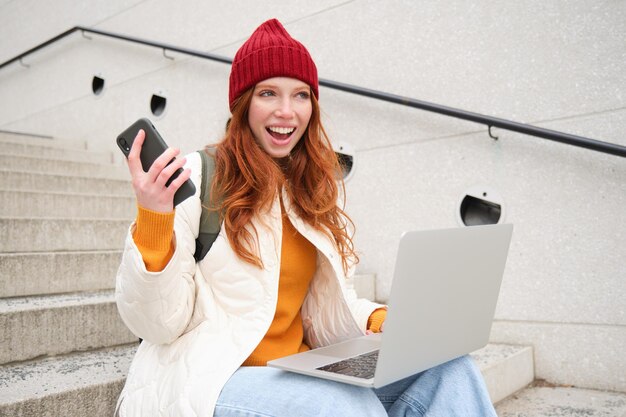 Femme rousse souriante avec téléphone portable et ordinateur portable assis dans les escaliers à l'extérieur du bâtiment se connecte à pu