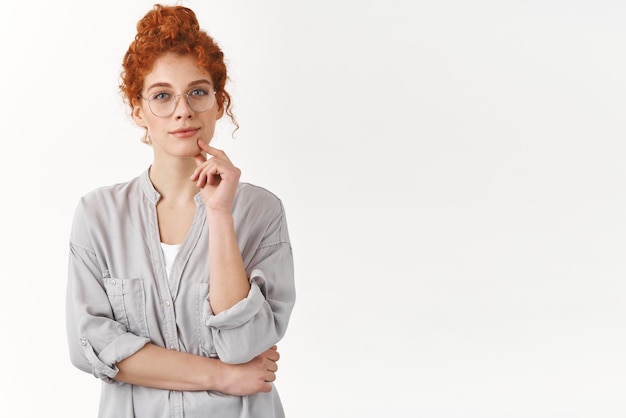 Femme rousse tendre et intelligente aux cheveux bouclés peignés en chignon regard avec un charmant sourire caméra toucher le menton pensif heureux de regarder la performance des enfants mignons debout fond blanc joyeux