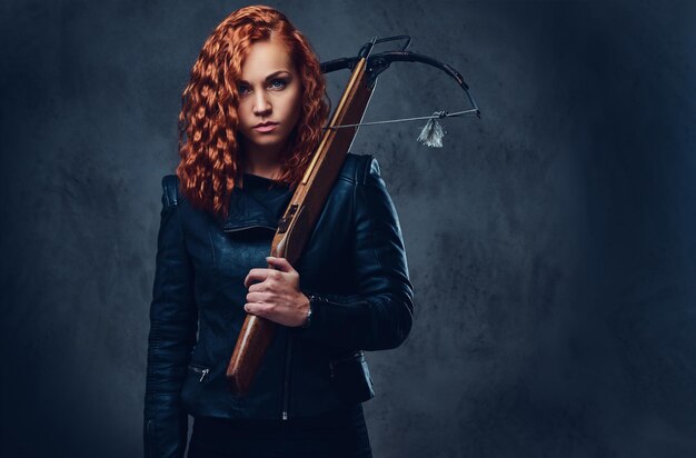 Une femme rousse vêtue d'un costume élégant tient une arbalète.