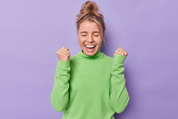 la femme s'exclame fort serre les poings se réjouit du succès sent le triomphe porte un col roulé vert isolé sur violet. un modèle féminin heureux célèbre la victoire