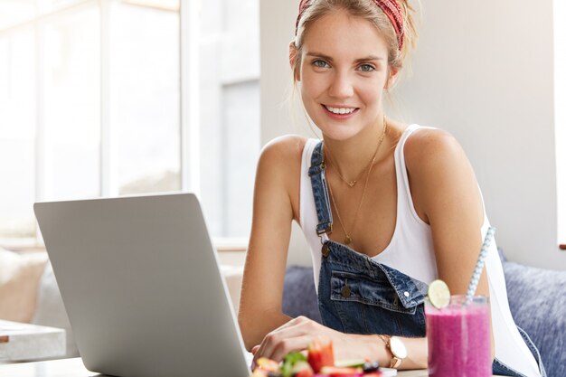 Femme en salopette en denim élégant avec ordinateur portable dans un café