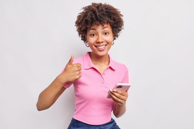 Une femme satisfaite tient un téléphone portable comme si le geste garde le pouce levé dit oui et recommande une nouvelle application vêtue de vêtements décontractés isolés sur un mur blanc