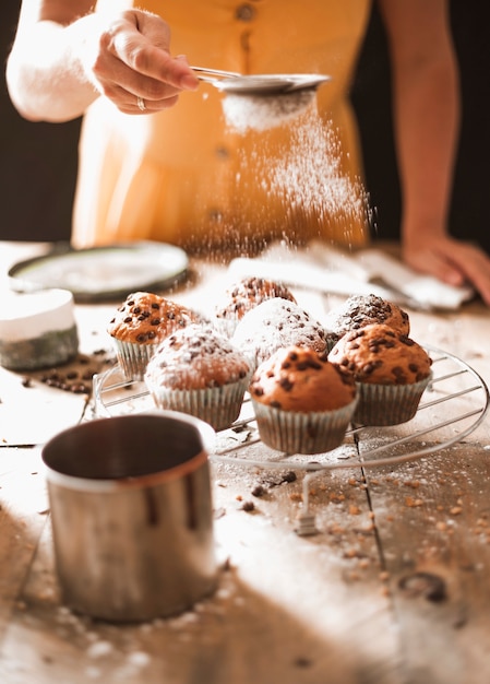 Une femme saupoudrant de sucre sur des muffins faits maison sur une grille de refroidissement